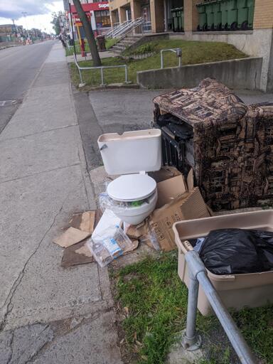 Toilet full of garbage on the side of the road next to an overturned chair