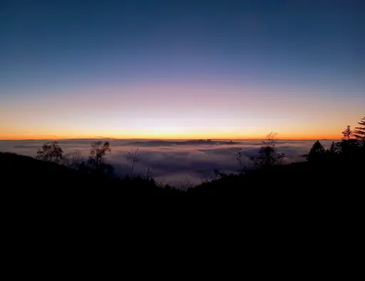 German: Ein majestätischer Abendhimmel über einem nebelverhüllten Tal, wobei sich in der Bildmitte sanft ein dunkler Bergrücken durch die Nebeldecke schiebt. Der Himmel zeigt einen perfekten Verlauf von tiefem Marineblau über zarte Violett- und Rosatöne bis hin zu einem intensiven Orange-Gelb am Horizont. Im Vordergrund zeichnen sich die schwarzen Silhouetten von Fichten und kahlen Birken ab. Das Nebelmeer nimmt die violetten Töne des Abendhimmels auf und umspielt den sich erhebenden Bergrücken wie eine sanfte Brandung.  English: A majestic evening sky over a fog-shrouded valley, with a dark mountain ridge gently pushing through the blanket of fog in the center of the image. The sky displays a perfect gradient from deep navy blue through delicate purple and pink tones to an intense orange-yellow at the horizon. In the foreground, black silhouettes of spruce trees and bare birch trees stand out. The sea of fog takes on the purple hues of the evening sky and swirls around the emerging mountain ridge like a gentle surf.