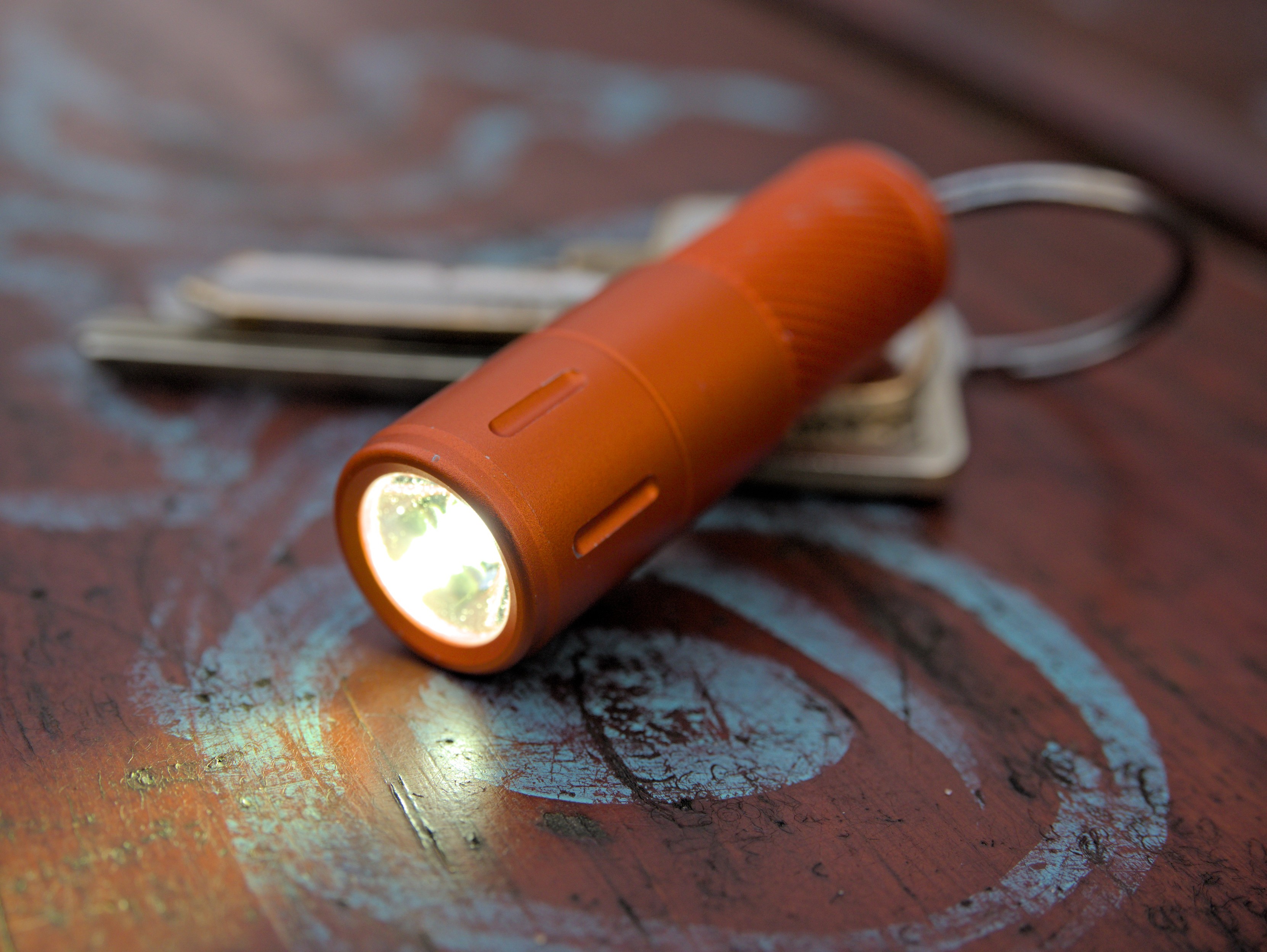 An orange Skilhunt EK1 flashlight attached to a keyring, turned on and illuminating a park bench